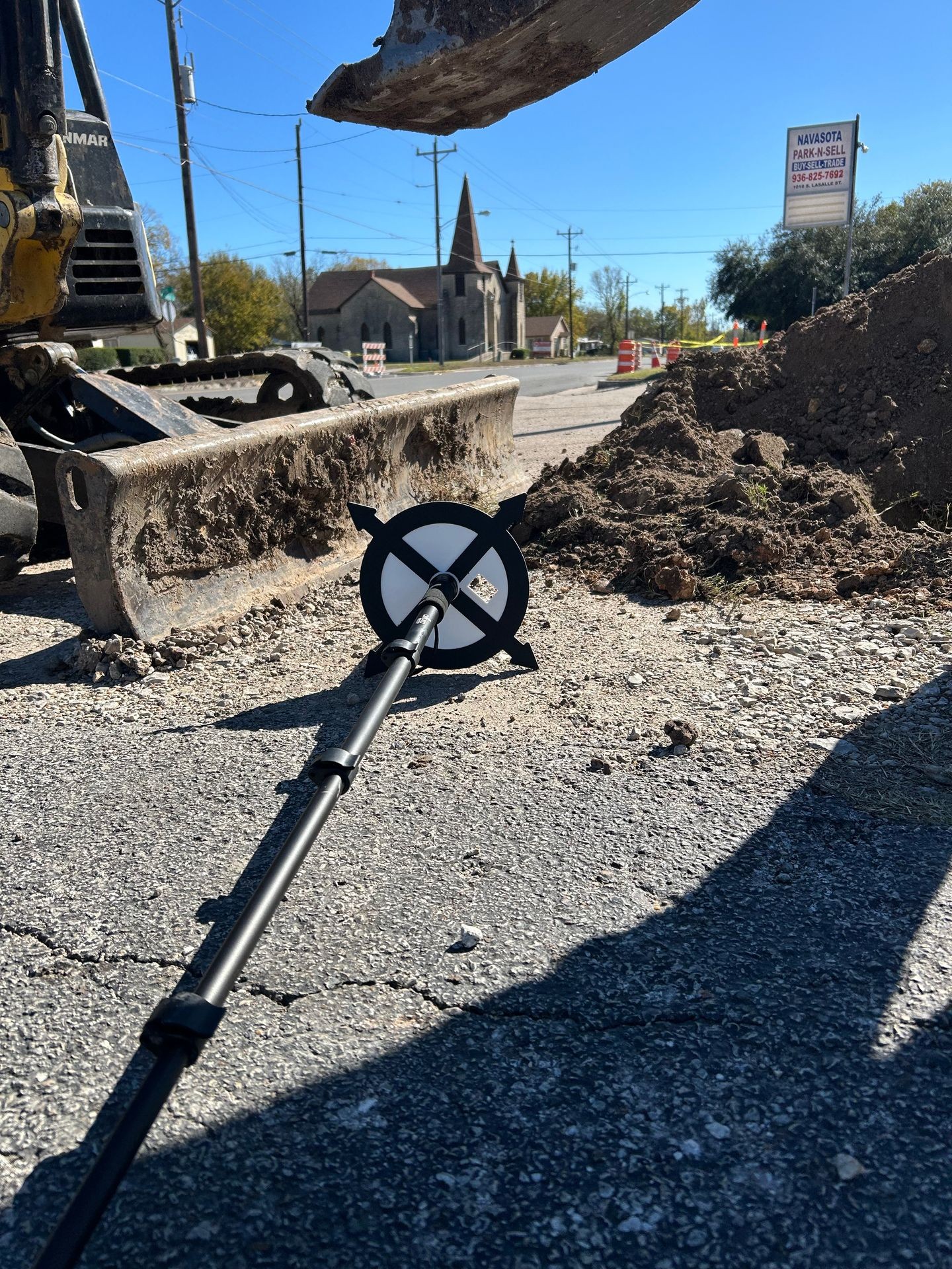 Construction site with excavator bucket, dirt piles, and a survey pole on a sunny day.